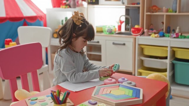 Girl stacking pastel wooden ring toys with bare hand on red table at playroom with pink chair and toy kitchen inside building; quiet concentration.