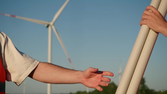 Construction engineer in white hard hat hands over technical documentation to field worker during renewable energy project. Close up of hand exchange, orange safety vest, and wind turbine blades in