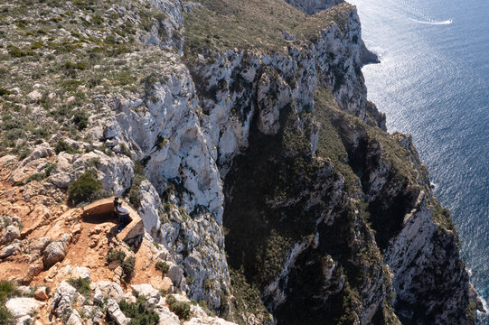 Aerial view of Mirador Josep Sastre lookout point on the edge of steep limestone cliffs with people looking at the blue sea in Andratx, Balearic Islands, Spain.