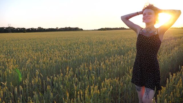 Cheerful punk girl in short black dress fooling around while walks among wheat meadow. Happy female hipster with tattoos having fun during goes through green barley field at sunset. Freedom concept