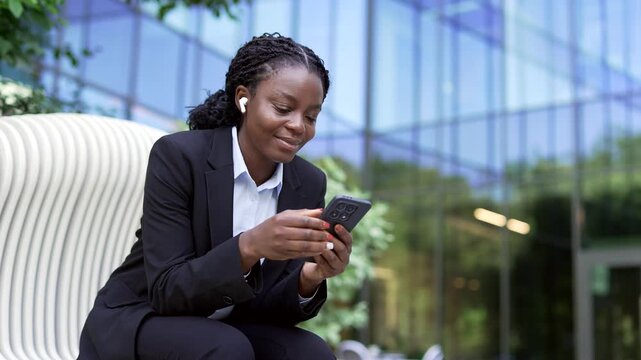 A happy african american businesswoman wears earphones and uses her smartphone while sitting on a bench near a glass building. A positive smiling corporate employee listening to a podcast or audio