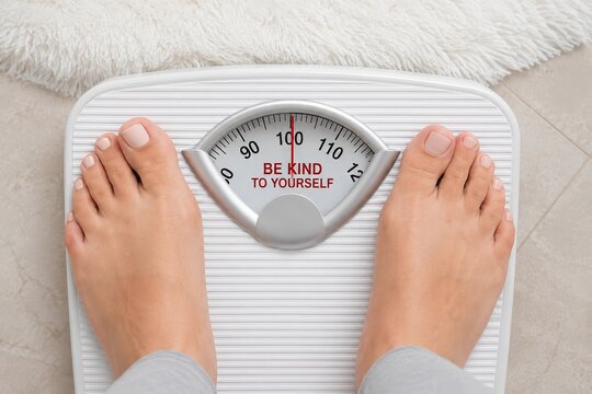 Feet on the scales (weight control).Top view of the legs of a woman standing on a scale, with the motivating inscription "be kind to yourself." weight, health, and self-esteem control.