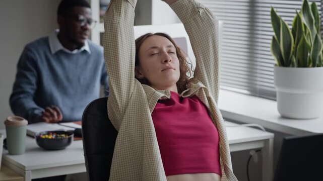 Medium tilt up shot of young adult Caucasian woman typing on computer and stretching arms overhead to relieve tension in office. For corporate wellness and office ergonomics marketing campaigns