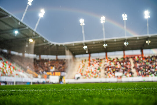 Soccer stadium illuminated by floodlights with rainbow over the roof during evening match, crowd watching game, real authentic sports scene.