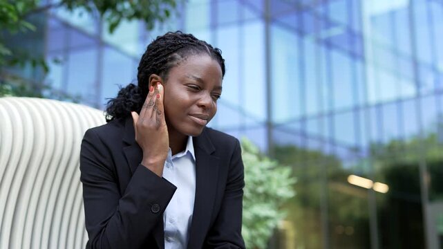 Close-up of a sick African American businesswoman in a suit suffering from an earache near an office building. Touching her ear with fingers on a bench, the female employee looks exhausted.
