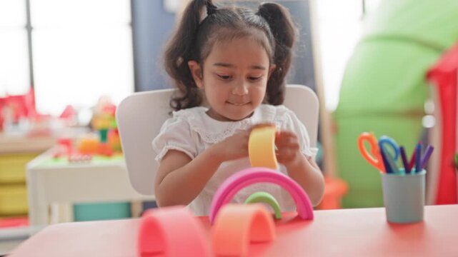 Little hispanic girl stacks colorful wooden rainbow toy with hands on table in building; quiet concentration.
