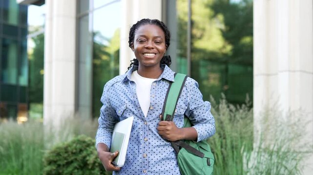 Portrait of a happy african american female student with backpack holding books and textbooks while standing on a university campus near a modern building. Smiling woman posing and looking at camera