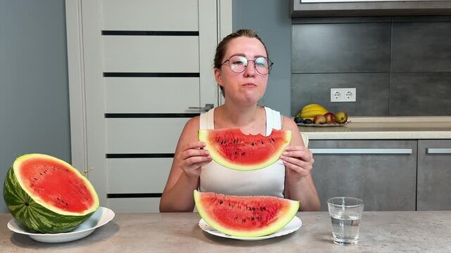 Middle-aged woman eating a large watermelon slice at a kitchen island