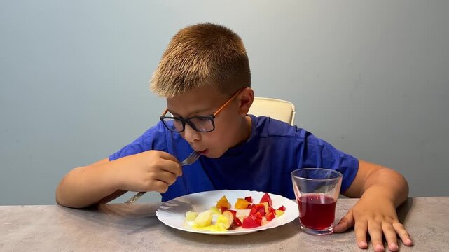 Young boy eating cut fruit at kitchen table with glass of red drink