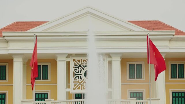 Vietnam Red flags fluttering past central fountain at neoclassical government in Ha noi building facade in public square.