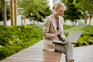 Businesswoman works on laptop in park on sunny day while holding glasses in hand