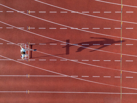 Aerial view of an athlete jumping over a hurdle on a red running track with a long shadow cast on the lanes in Almere, Flevoland, Netherlands.