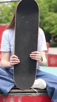 Girl sitting in skate park, holding skateboard in front of her. Front view of deck with black griptape. Teenager resting after skateboarding. Concept of active recreation, street culture and youth lei