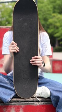Girl sitting in skate park, holding skateboard in front of her. Front view of deck with black griptape. Teenager resting after skateboarding. Concept of active recreation, street culture and youth lei