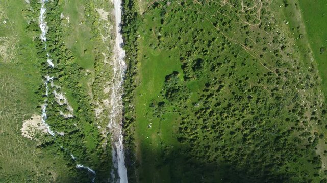 Birds eye view drone shot of a gravel road and winding mountain river stream running through lush green bushes and meadows in Sairam-Ugam National Park, Kazakhstan. 