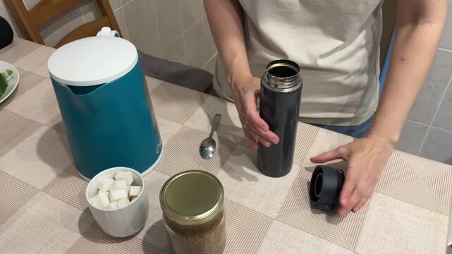 white woman filling thermos with coffee at kitchen table scooping instant coffee from glass jar and adding sugar cubes closeup of hands calm warm light cozy morning ritual home barista vibe