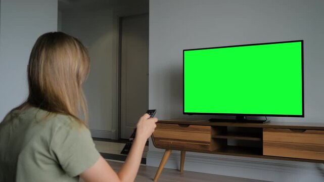Woman is watching flat smart led TV with blank green display and changing the channels with remote in grey living room at home. Technology, mock up, template, chroma key, and green screen concept