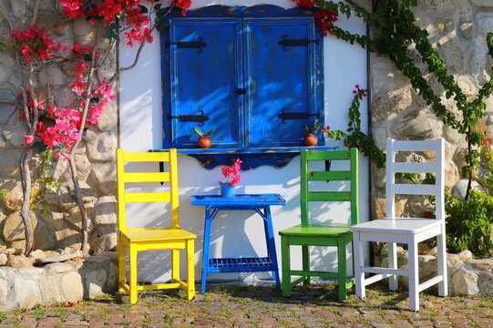 A street cafe in a resort town. The wall of a white house, blue shutters of a cafe, and colorful chairs and a small table i