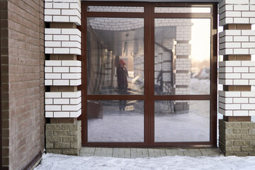 Naklejka premium Glass Entrance Door Reflecting Person And Snowy Courtyard At Building Facade With White Brick Columns And Tiled Threshold During Cold Daylight.
