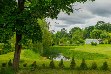 Peaceful Latvian farm landscape with pond and lush greenery under dramatic summer cloudy sky scene © Zigmunds