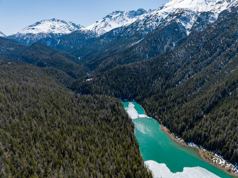Aerial view of a turquoise alpine lake surrounded by dense evergreen forests and snow-capped mountain peaks near Fuorn Pass in Val Mustair, Grisons, Switzerland.
