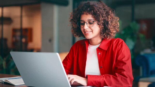 Young indian woman with curly hair and glasses working on laptop computer at desk in modern office or home workspace, wearing red shirt and smiling. Latin or muslim arab student girl studying indoors