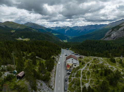Aerial view of Fuorn Pass featuring a mountain road, a hotel building, and dense pine forests under a cloudy sky in Val Mustair, Grisons, Switzerland.