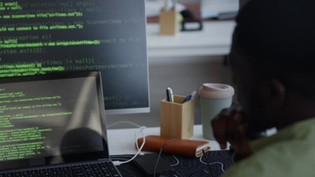 Close up of young adult African American man analyzing programming code on computer screens in office. Clip ideal for technology, software development, and cybersecurity marketing campaigns