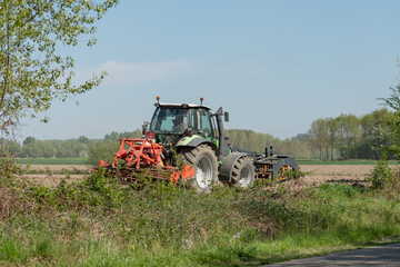 Fototapeta premium Diepenbeek, Belgium. Tractor cultivating agricultural field next to rural roadside with vegetation and trees