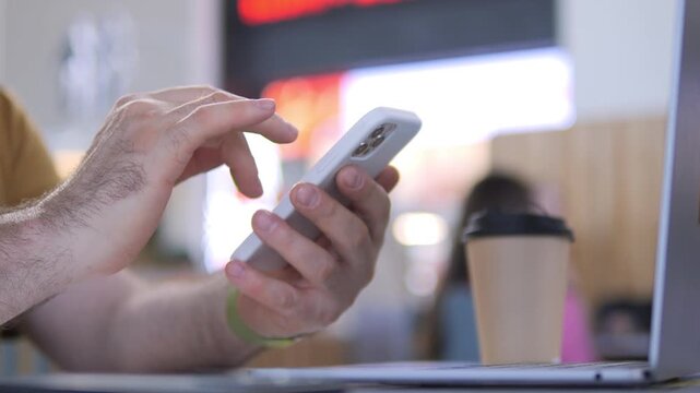 Hand scrolls smartphone while laptop and coffee cup sit nearby. Blurred background suggests busy cafe or office setting. Person focused on phone, multitasking with tech devices