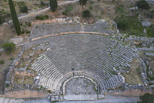 Aerial view of the ancient theatre of Delphi featuring its well-preserved stone tiers and semicircular orchestra nestled on a rocky hillside with cypress trees in Delphi, Greece.