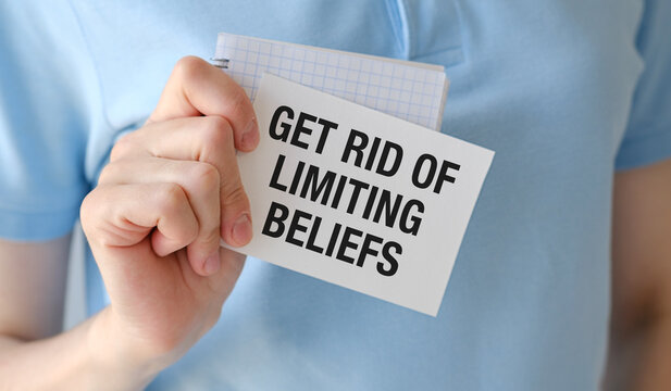 Hand holds note with bold message: Get rid of limiting beliefs. Person wears soft blue shirt, focused and determined.