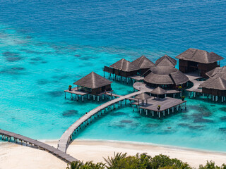 Fototapeta premium Aerial view of a Maldives atoll resort shows a curving boardwalk, thatched overwater villas, white sand, and lagoon gradients under midday sun and clear skies.