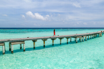 Fototapeta premium A wooden jetty in the Maldives extends over turquoise shallows to deep water, with one person in a bright red dress walking, under midday sun and soft clouds.