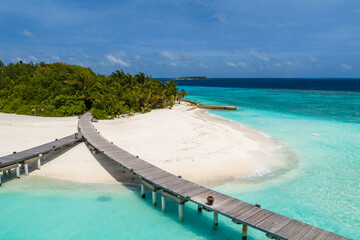 Fototapeta premium Aerial view of a Maldives resort island with a wooden boardwalk over turquoise lagoons, a crescent of white sand, green palms, and a jetty in bright daylight.