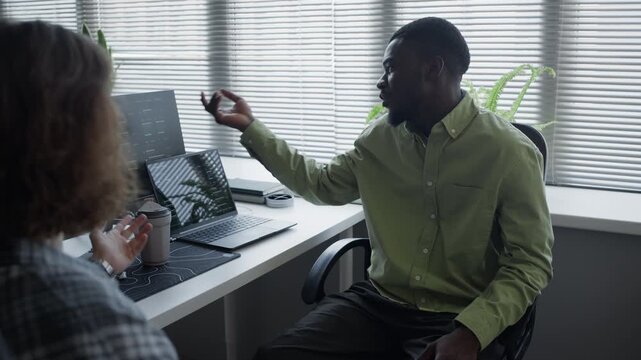 African American man and Caucasian colleague discussing software development at office desk with computer screens, slow motion. Footage for tech industry marketing and corporate training videos