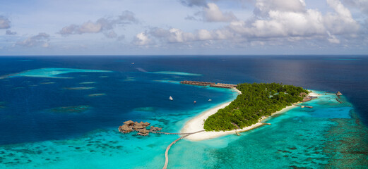 Obraz premium Aerial scene of a Maldives resort island with white sand, turquoise shallows, overwater villas on a curved pier, wooden pavilions, jetty, palms, and midday light.
