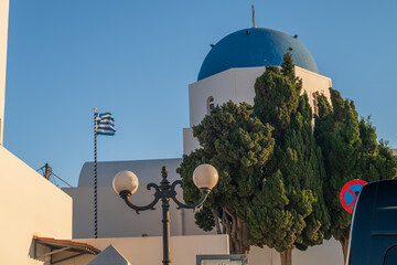 Whitewashed Cycladic church with blue dome, Greek flag, and streetlamp under clear summer sky on Santorini island, Greece. © Andrei Tomas