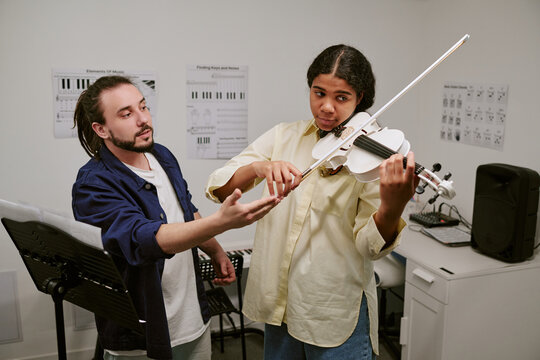 Teenager playing violin while male instructor guiding bow technique during music lesson, medium shot. Useful for education marketing, arts training, private tutoring