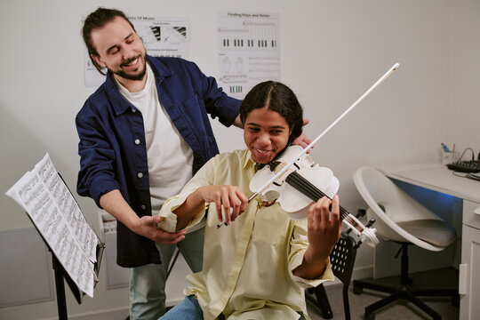 Teenage girl playing violin during lesson while male instructor guiding posture and bow technique in music classroom. Ideal for education marketing, arts programs, private tutoring
