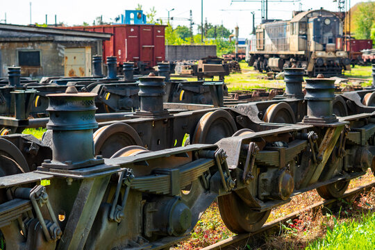 Close-up of steel railway wagon bogies with leaf springs and wheels on tracks at a train depot; a blurred background features an old diesel locomotive, a red freight car, and station infrastructure in