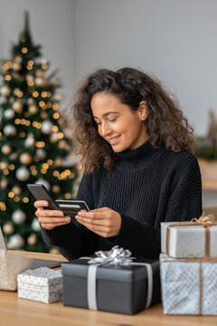 Young woman with curly hair in black sweater checks her smartphone while sitting at a table surrounded by Christmas gifts and a decorated tree in the background