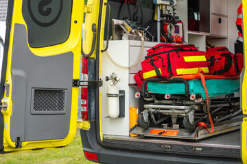 Interior of a modern yellow emergency ambulance vehicle with open rear doors, featuring professional medical stretcher with red trauma bags, oxygen tank, resuscitation equipment, and medical details i © Adam