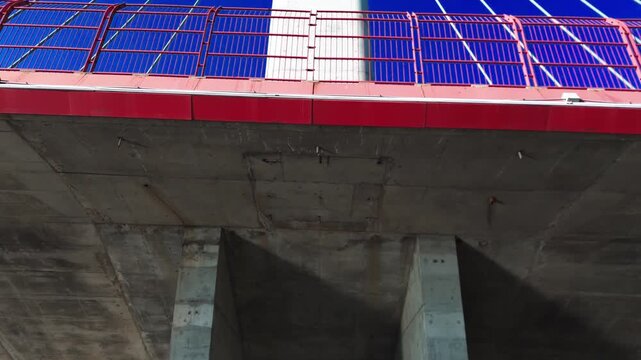 Concrete bridge underside fills the frame. Red railings and blue cables stretch into the distance. Sunlight creates deep shadows on the structure. Solar panels cover the bridge deck