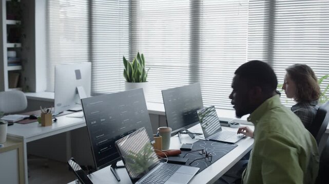 Tracking shot of African American young adult man sitting down at office desk to write code on laptop next to Caucasian female colleague. For promoting IT startups and digital workspace solutions