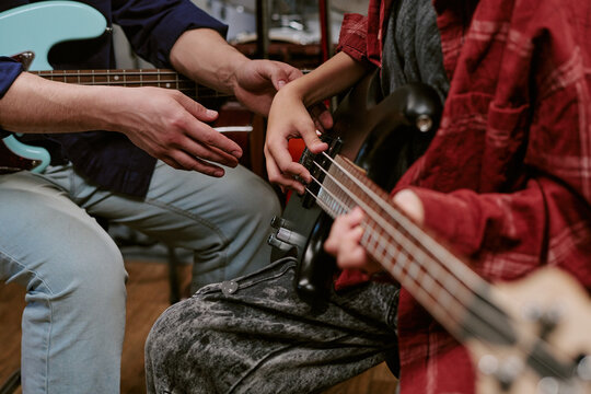 Close up of teenager learning bass guitar with adult instructor guiding hand placement during music lesson, showing skill development and mentoring for education marketing