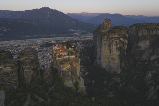 Aerial view of the Holy Monastery of Varlaam perched on a massive sandstone rock pillar at dusk with the town of Kalabaka and distant mountains in the background in Trikala, Greece.