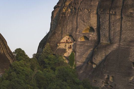 Aerial view of the historic Meteora monastery built into the side of a massive sandstone cliff surrounded by lush green forest in Trikala, Greece.