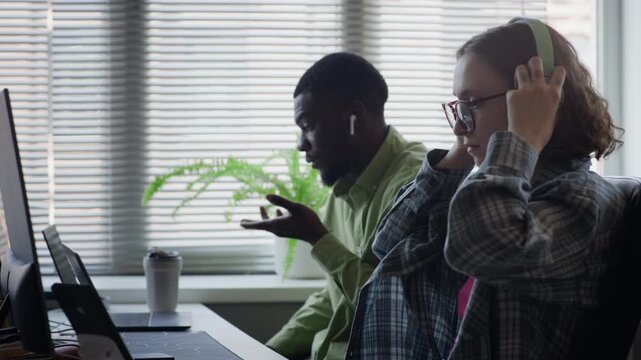 Side view of Caucasian young adult woman putting on headphones and typing on laptop next to African American young adult man working on computer. Perfect for software development or tech startup promo