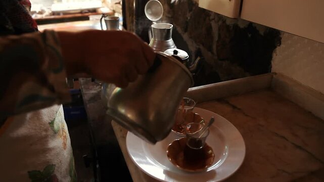 Pouring steaming hot traditional Turkish tea from a metal teapot into classic tulip-shaped glasses.